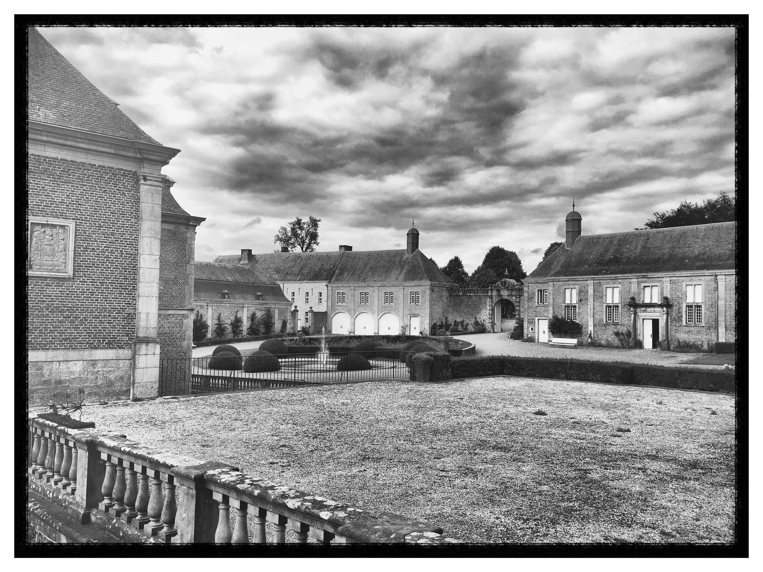 A historic courtyard with brick buildings, a central fountain, and a large tree in the background.