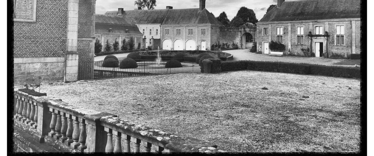 A historic courtyard with brick buildings, a central fountain, and a large tree in the background.