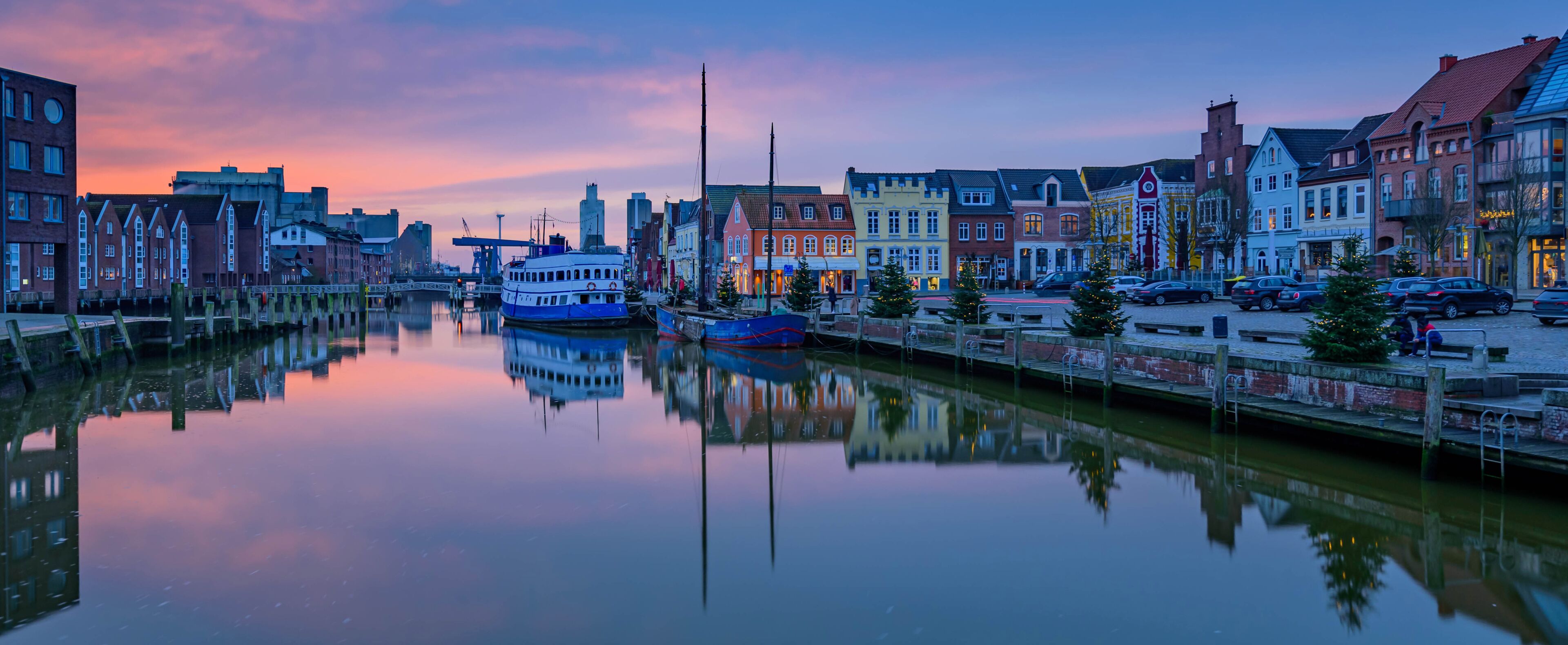 A calm river reflecting buildings, a blue boat, and a sunset sky.