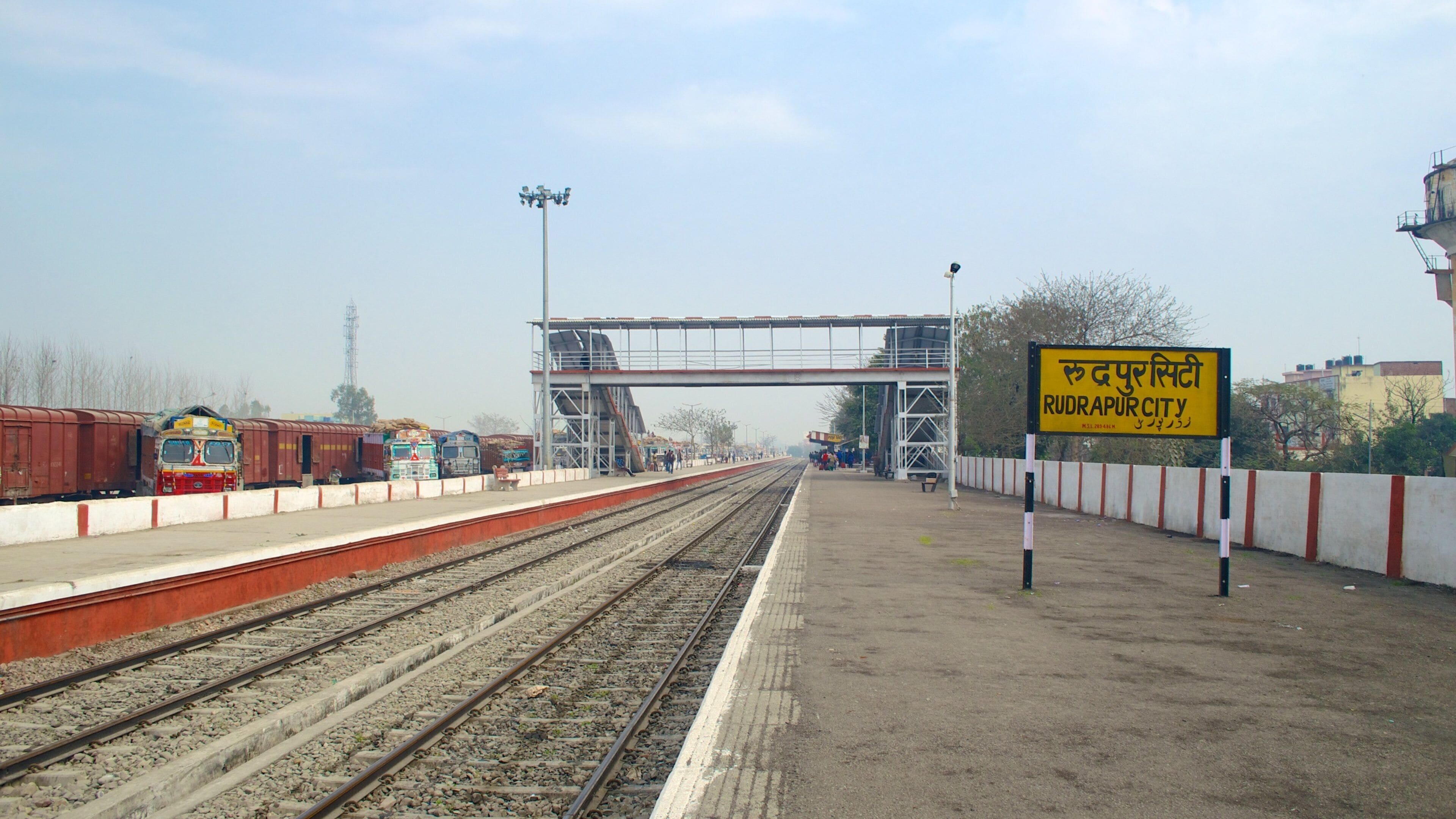 A railway station platform with multiple tracks, a pedestrian overpass, and a sign indicating 'RUDRAPUR CITY'.