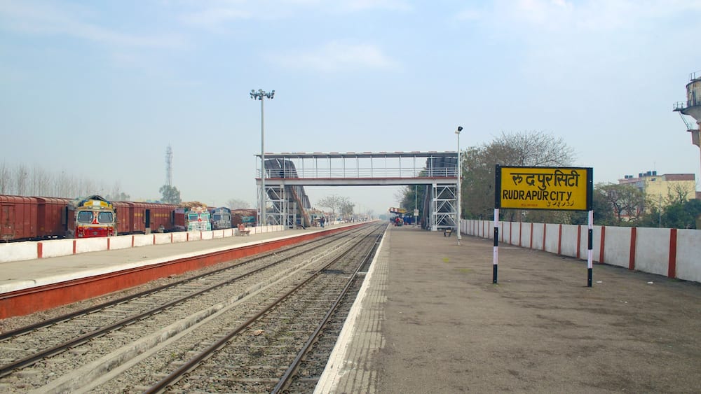 A railway station platform with multiple tracks, a pedestrian overpass, and a sign indicating 'RUDRAPUR CITY'.
