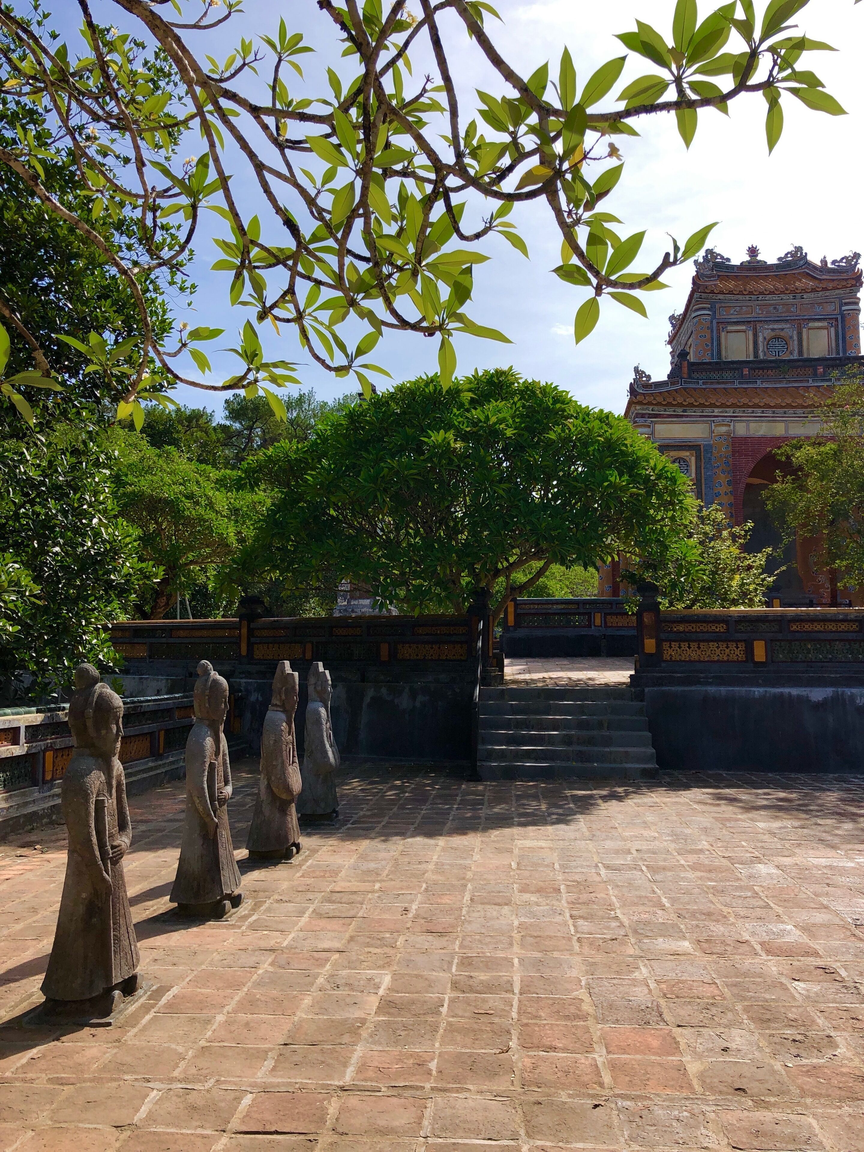A courtyard with stone statues, a large tree, and a traditional building in the background.