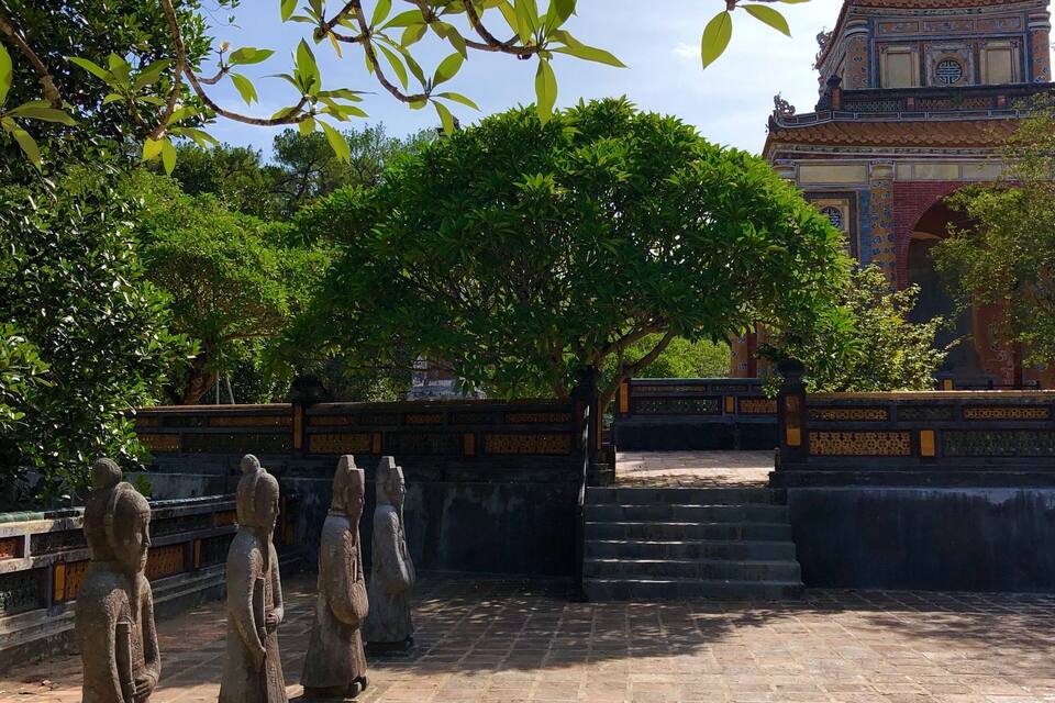 A courtyard with stone statues, a large tree, and a traditional building in the background.