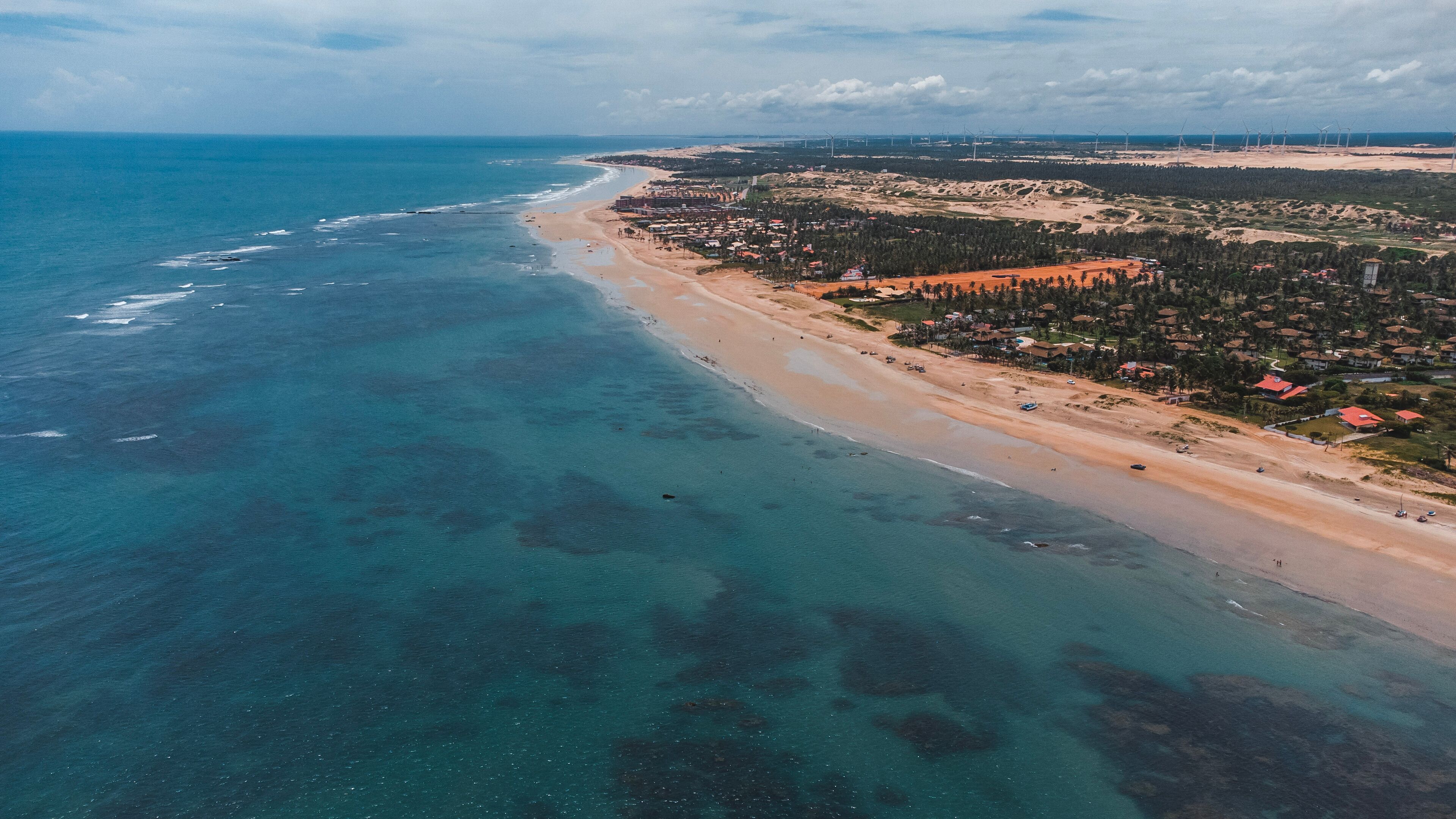 A coastal town with a beach, clear blue waters, and wind turbines in the background.