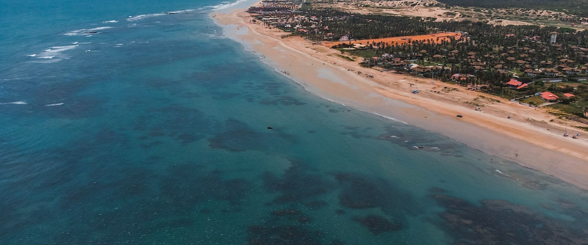 A coastal town with a beach, clear blue waters, and wind turbines in the background.
