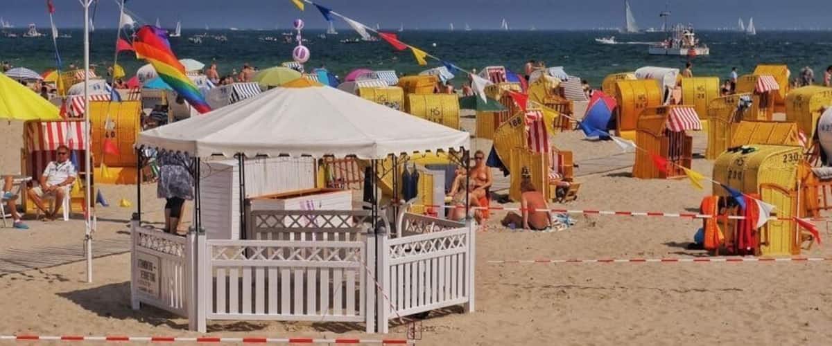 A crowded beach with colorful beach huts and a white kiosk.