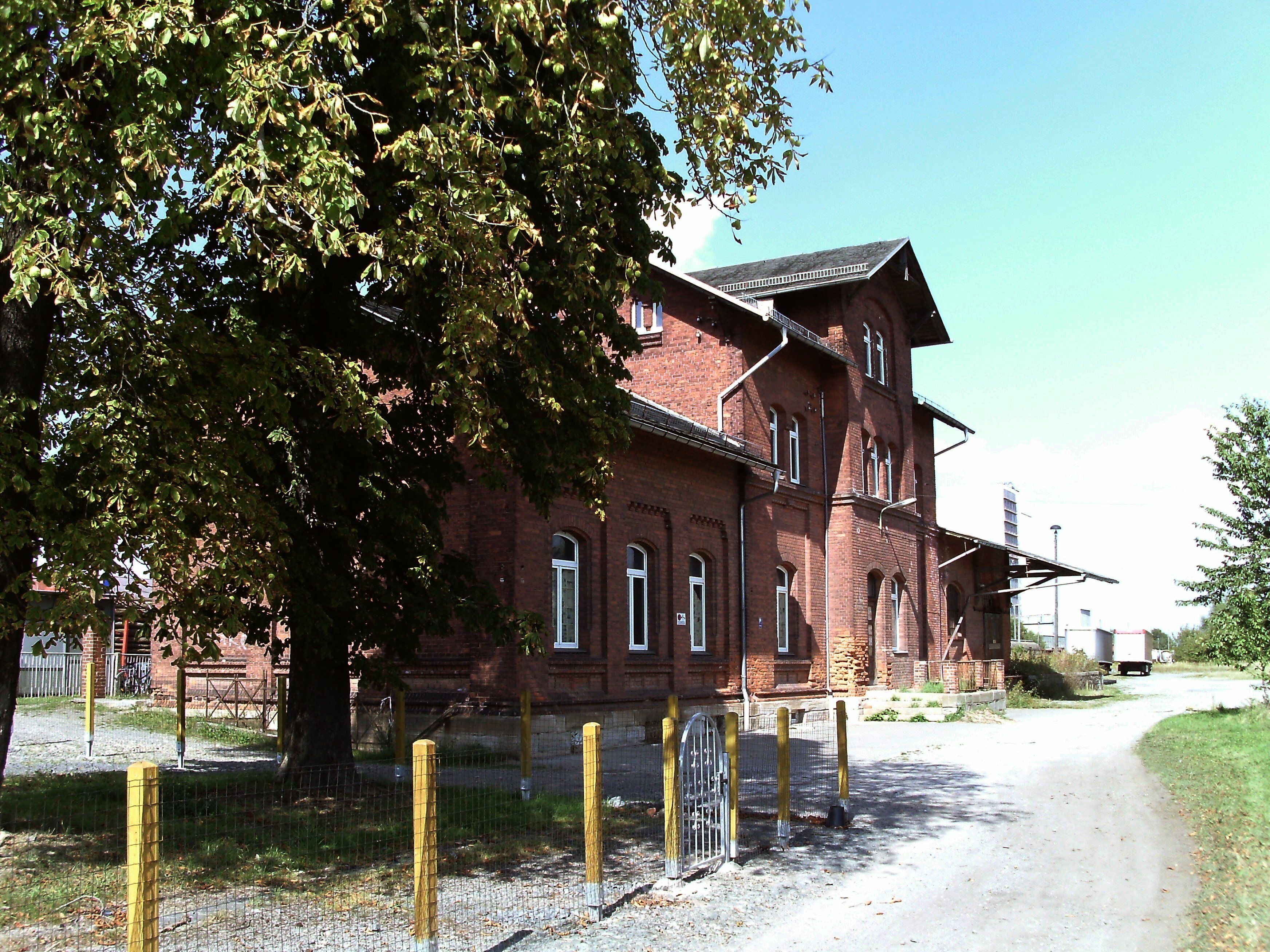 A large brick building with multiple windows and a sloped roof, surrounded by trees and a gravel path.
