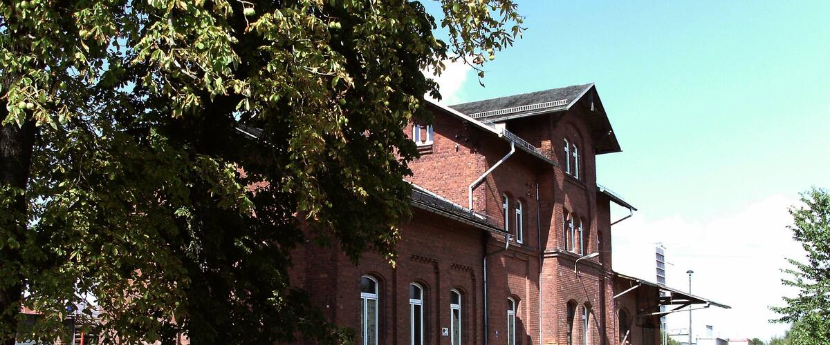 A large brick building with multiple windows and a sloped roof, surrounded by trees and a gravel path.