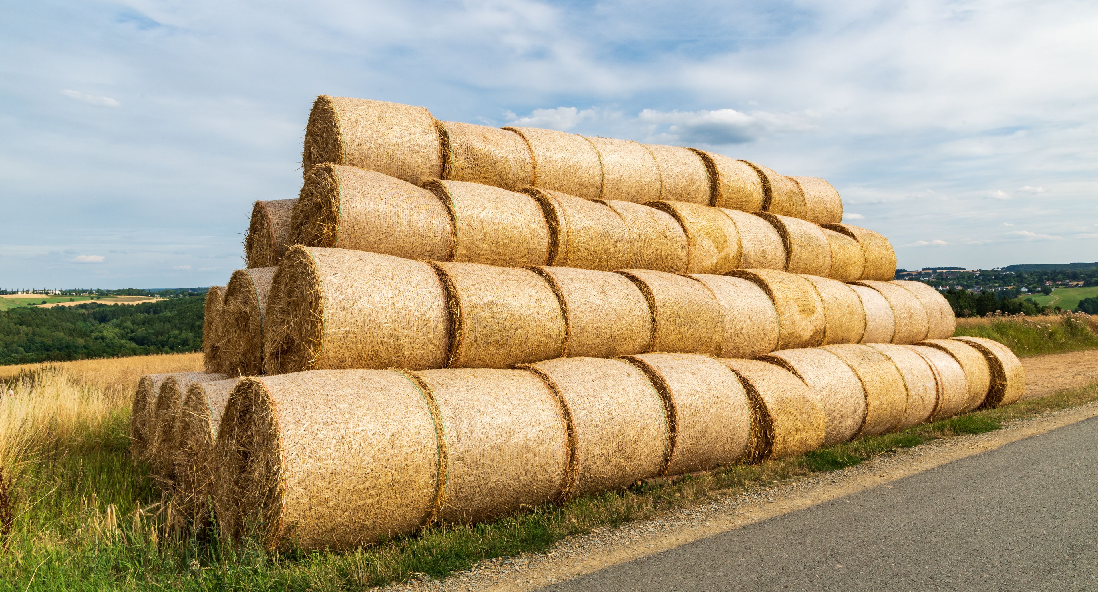 Large hay bales stacked on a roadside with a rural landscape in the background.