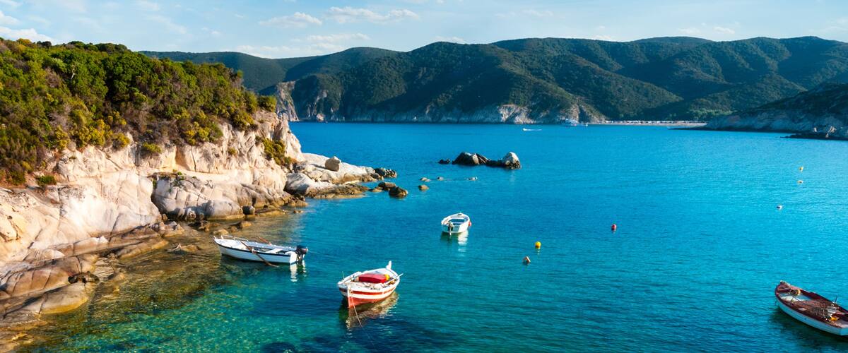 A coastal landscape with clear blue waters, rocky cliffs, and boats anchored near the shore.