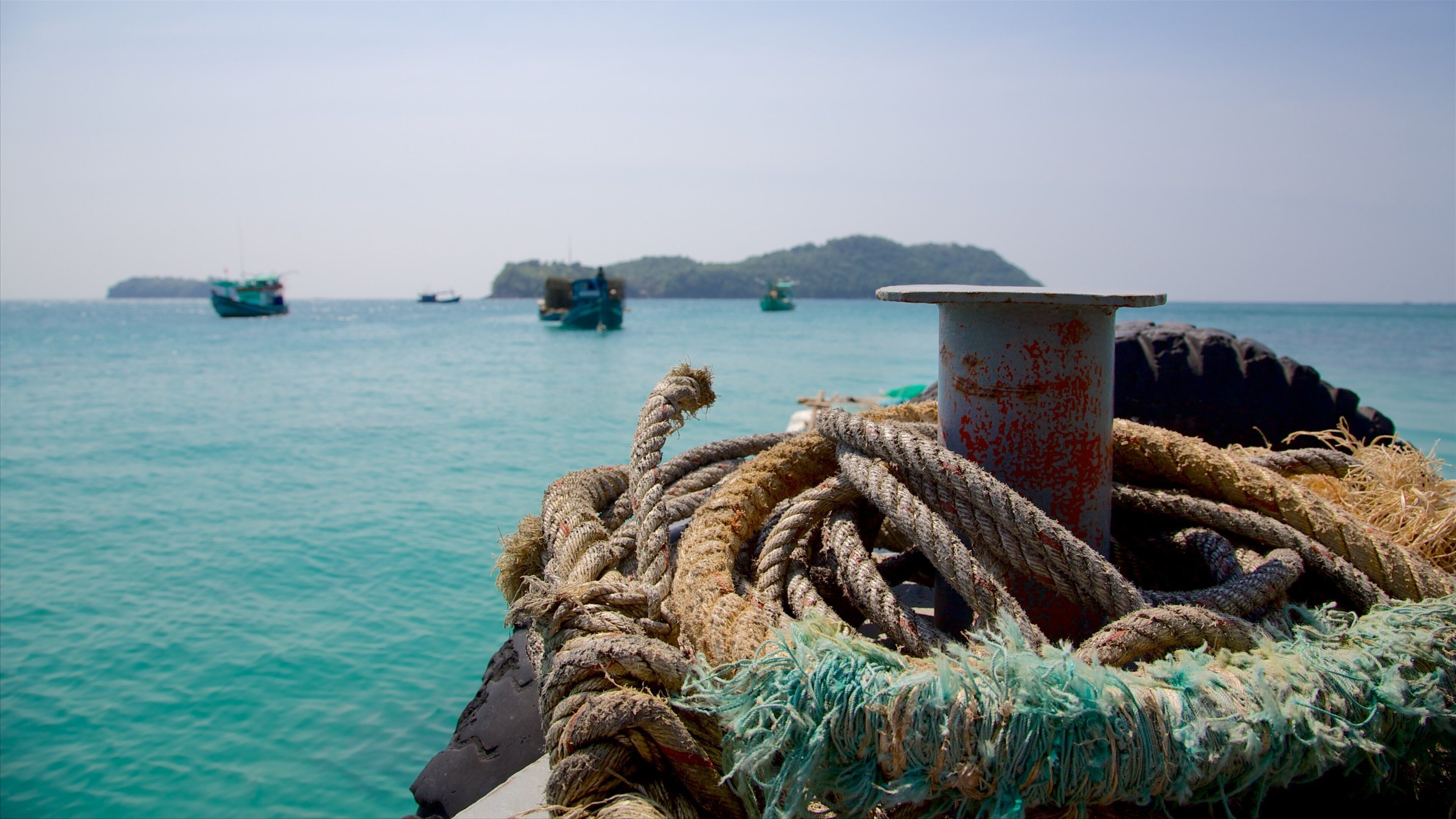 A boat's bow with ropes and a metal bollard, calm turquoise sea, and distant boats and land.