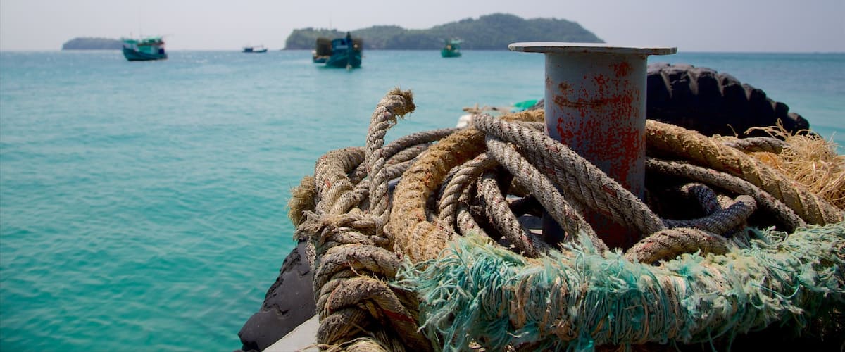 A boat's bow with ropes and a metal bollard, calm turquoise sea, and distant boats and land.