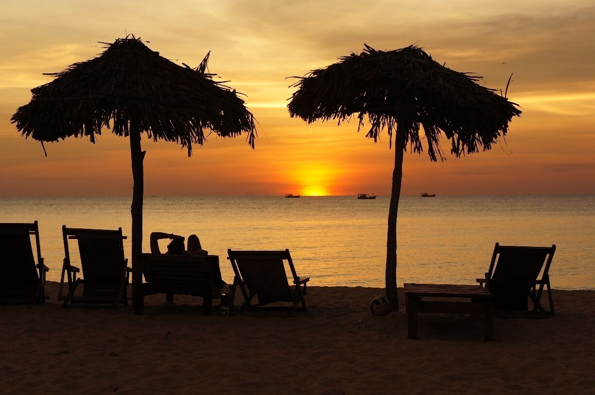 A beach with thatched umbrellas and wooden chairs at sunset.