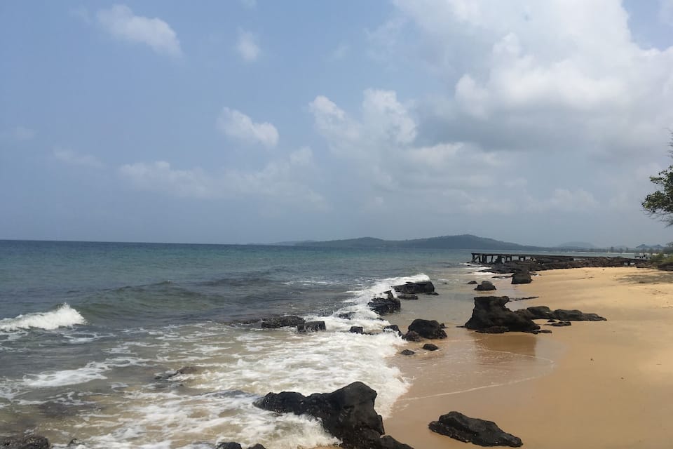 A rocky beach with waves, a wooden pier, and distant hills.