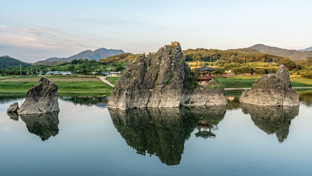 A calm lake with three prominent rock formations in the foreground, a traditional pavilion on the right, and a mountainous landscape in the background.