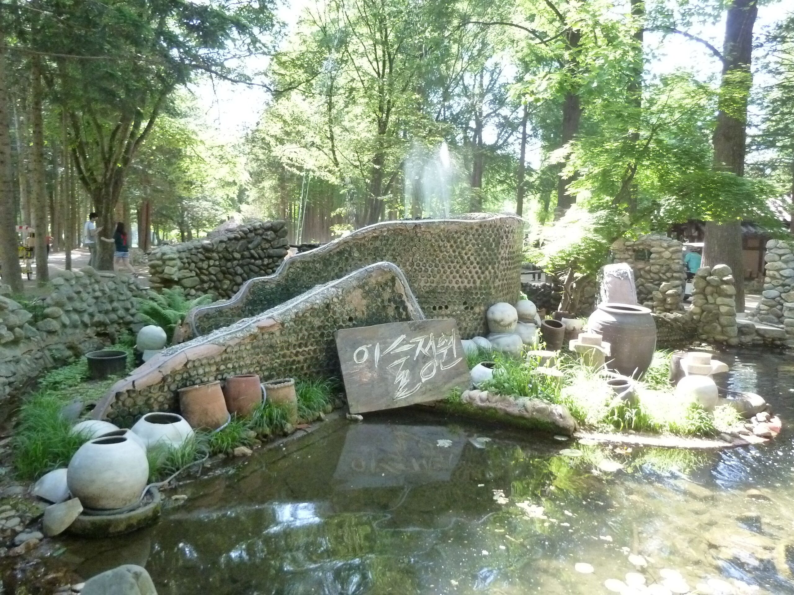 Un jardín tranquilo con un estanque, esculturas de piedra y un letrero con caracteres coreanos.