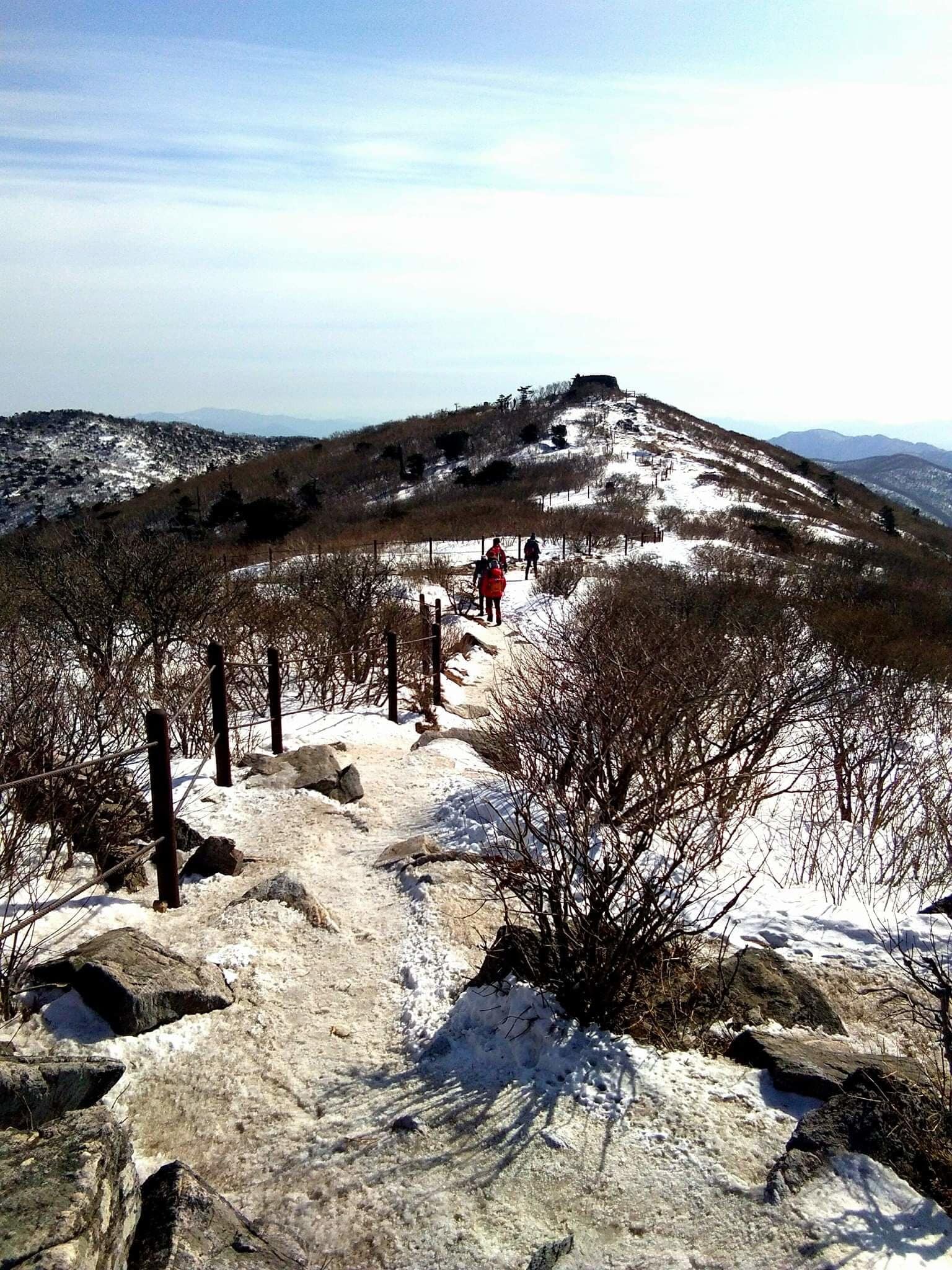 A snowy mountain trail with hikers, rocky terrain, and sparse vegetation.