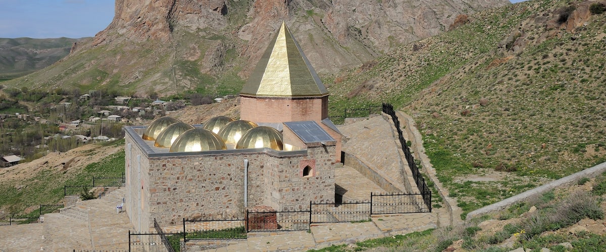 A stone building with a golden spire and domed roof, surrounded by a mountainous landscape.