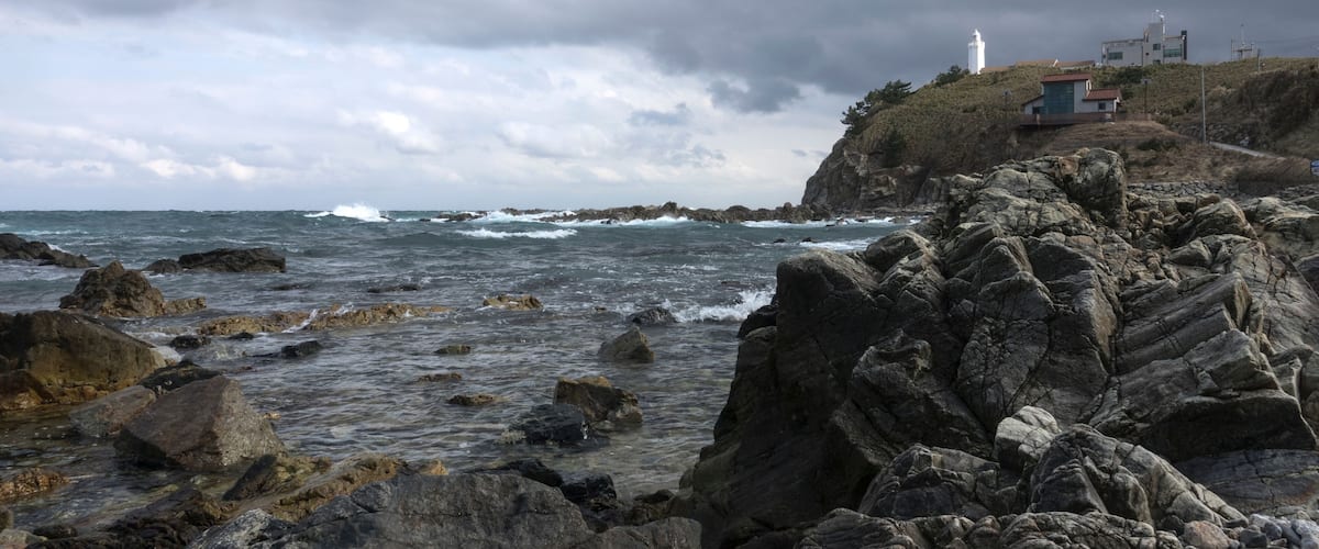 A rocky coastline with waves crashing against the shore, a lighthouse, and buildings on a hill.