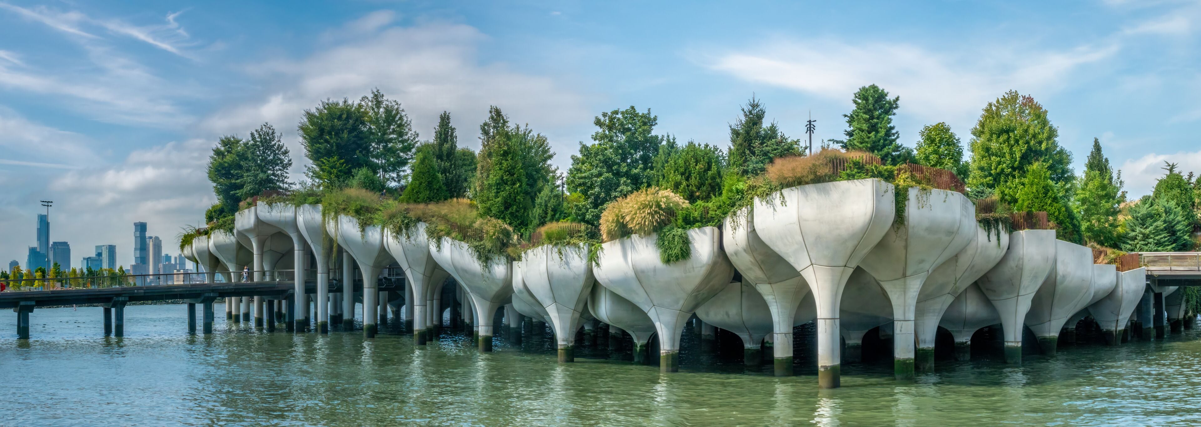 Un ponte originale caratterizzato da strutture organiche simili ad alberi, che lo sostengono su un corso d'acqua, con sullo sfondo lo skyline di una città.