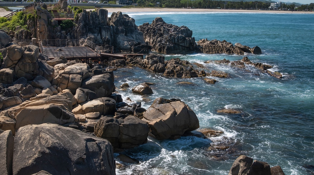 A rocky coastline with waves crashing against large boulders, a bridge, and a distant beach with mountains in the background.