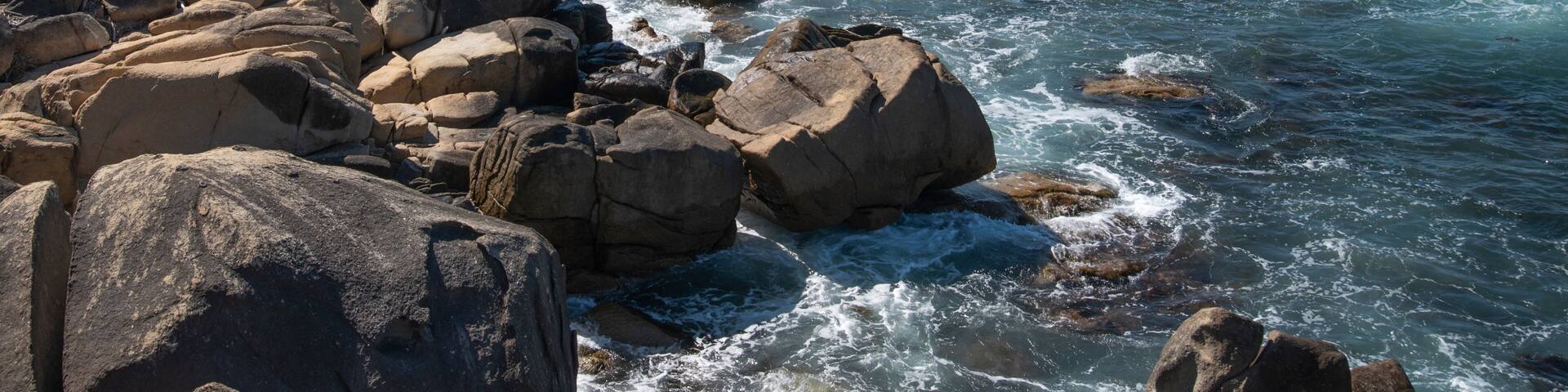 A rocky coastline with waves crashing against large boulders, a bridge, and a distant beach with mountains in the background.