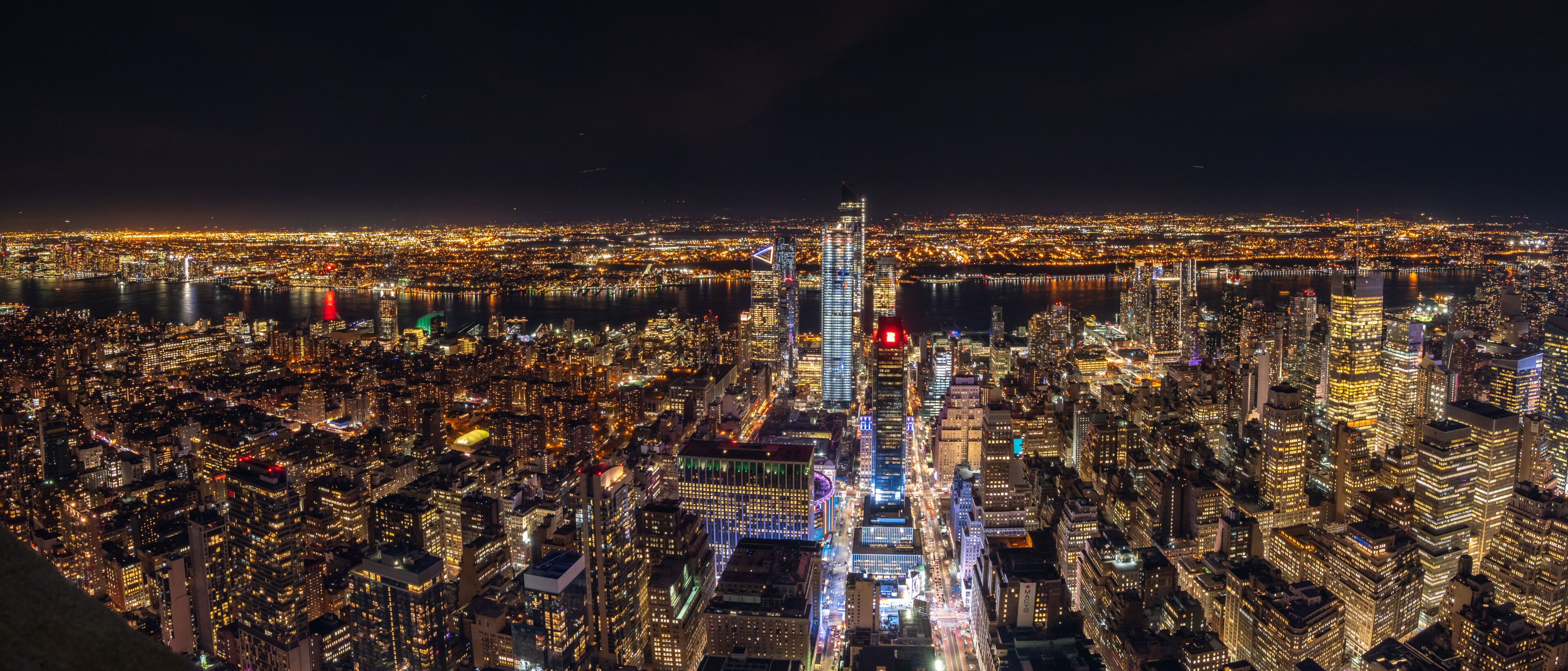 A nighttime cityscape with a prominent skyscraper, dense urban buildings, and a river.