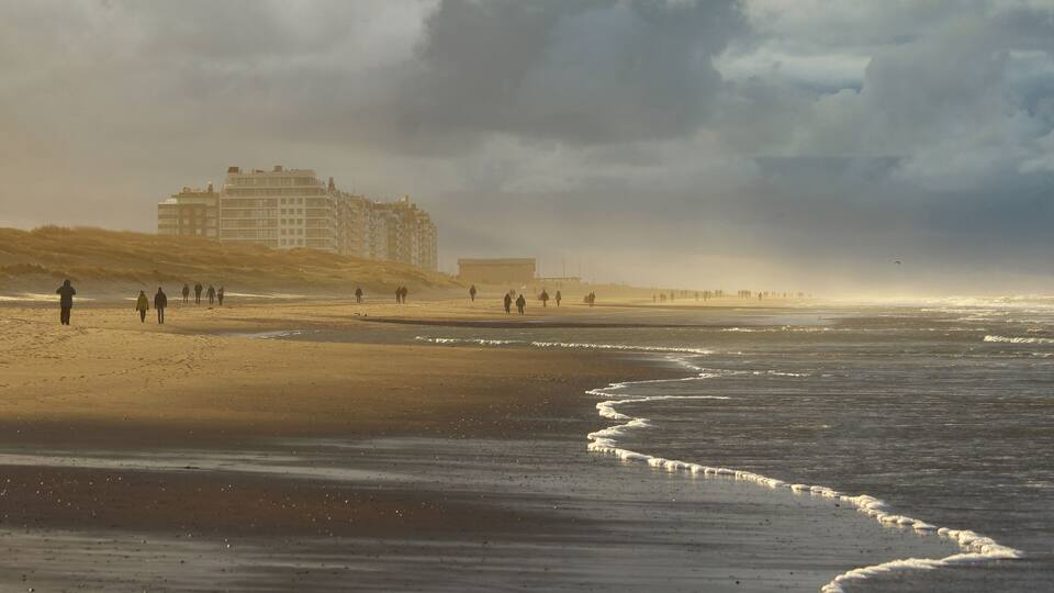 A beach with people walking, a large building in the background, and waves approaching the shore.