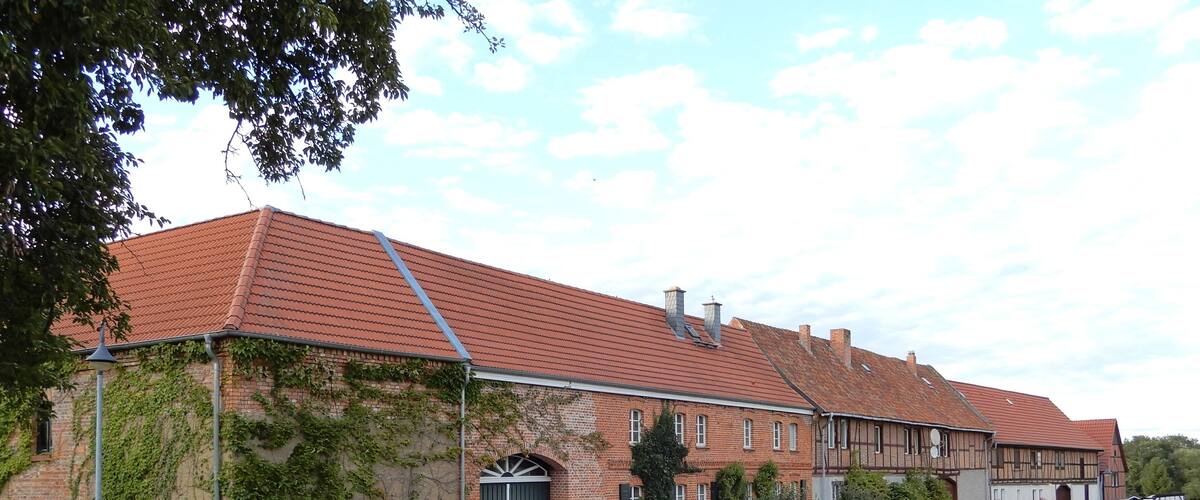 A large brick building with a red-tiled roof, surrounded by greenery and a clear sky.