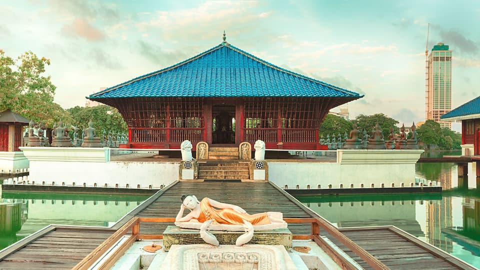 A traditional pavilion with a blue tiled roof, a statue of a reclining figure, and a calm water body reflecting the structures.