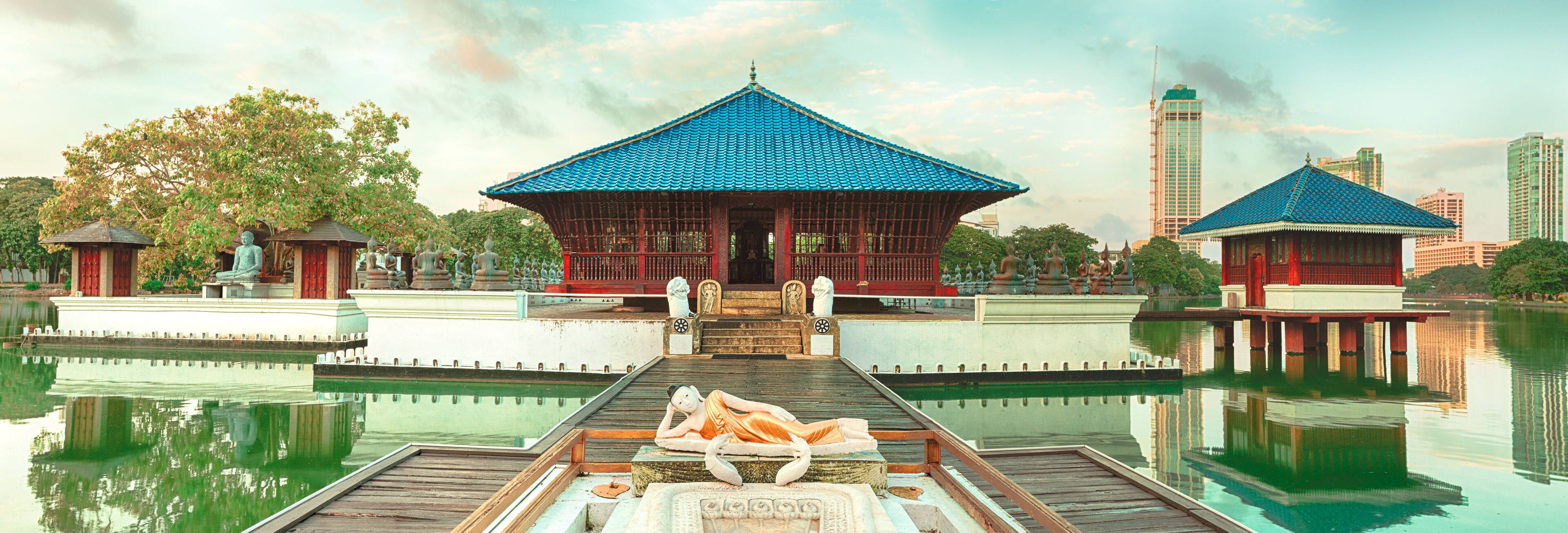A traditional pavilion with a blue tiled roof, a statue of a reclining figure, and a calm water body reflecting the structures.