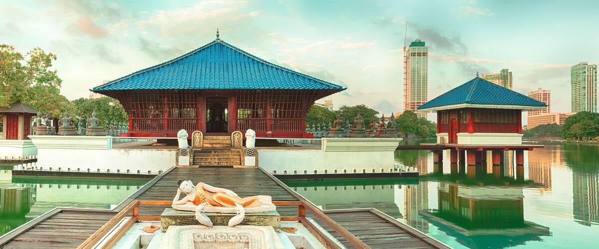 A traditional pavilion with a blue tiled roof, a statue of a reclining figure, and a calm water body reflecting the structures.