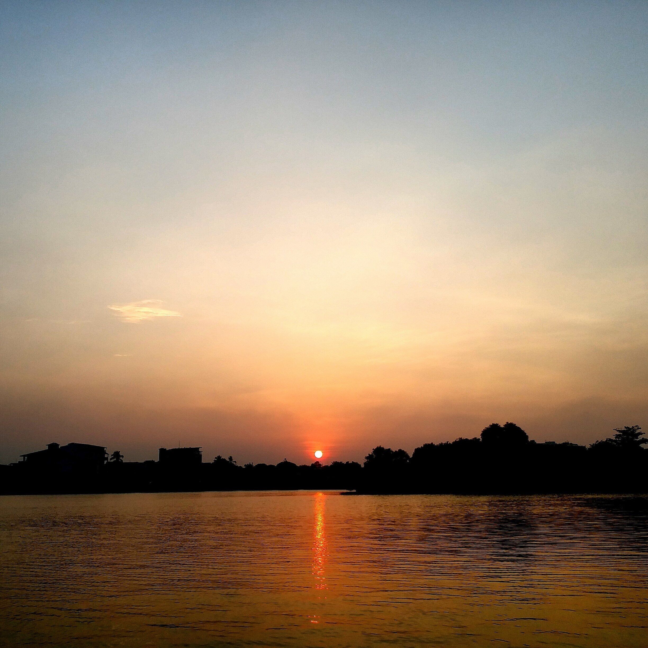 A sunset over a calm body of water with silhouettes of trees and buildings on the horizon.