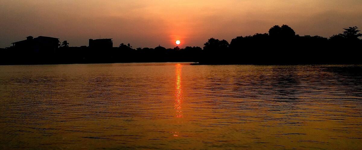 A sunset over a calm body of water with silhouettes of trees and buildings on the horizon.