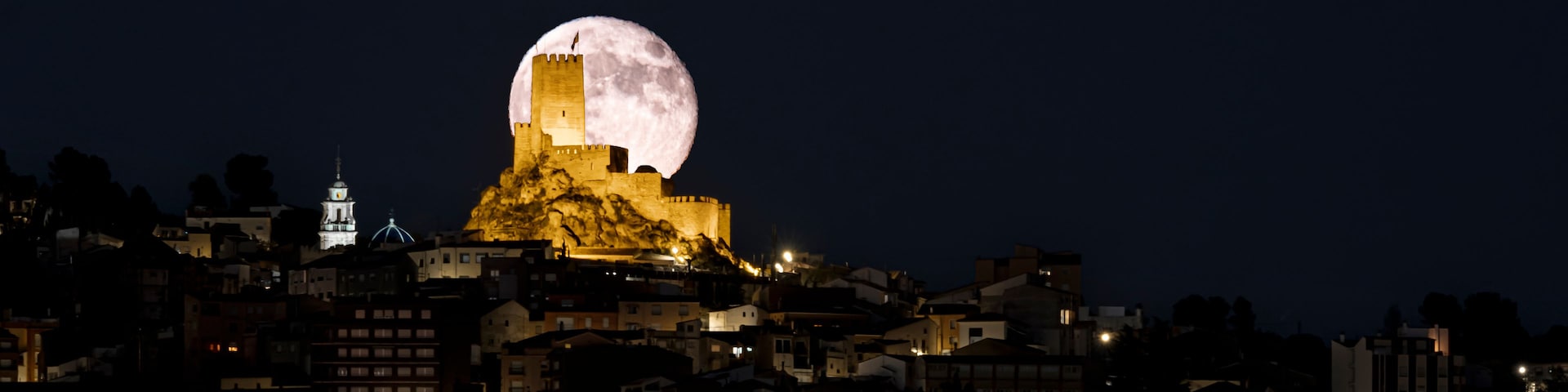 Paisagem urbana noturna com um castelo imponente numa colina, iluminado por luzes artificiais, e uma grande lua cheia no céu.