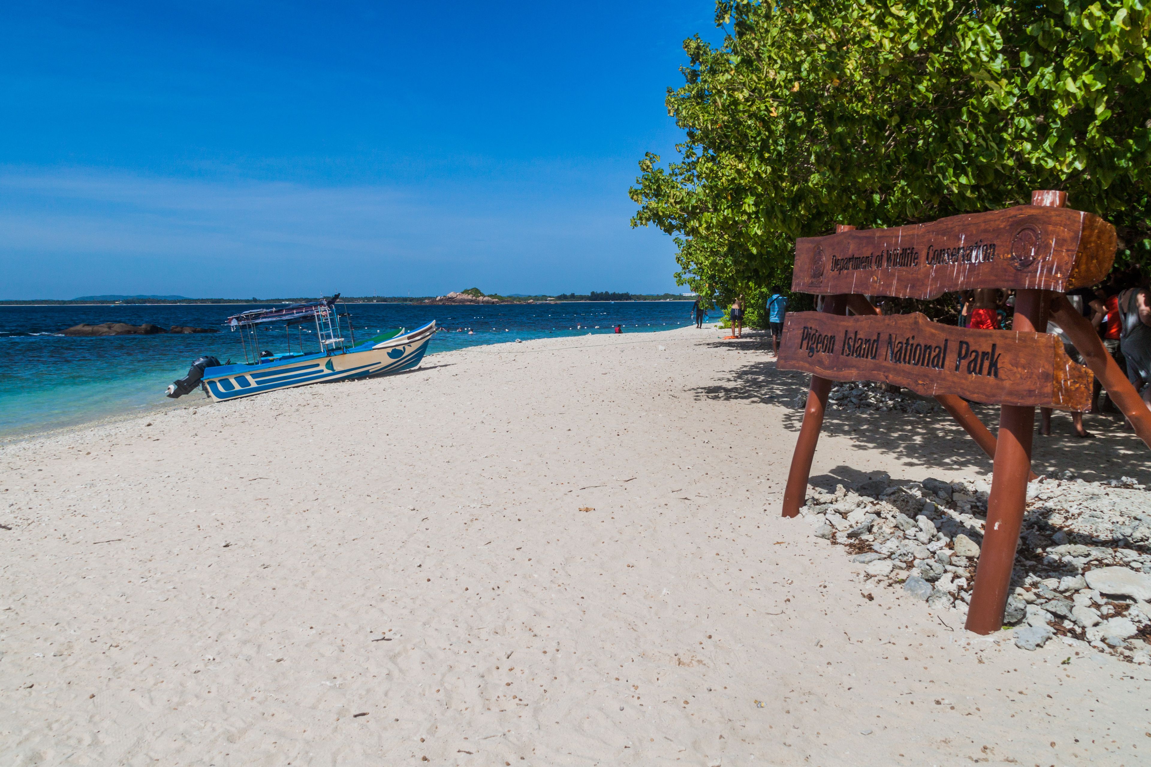 A beach with a wooden sign for Pigeon Island National Park, a boat on the shore, and clear blue water.