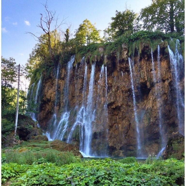 A multi-tiered waterfall cascading down a rocky cliff surrounded by lush greenery.