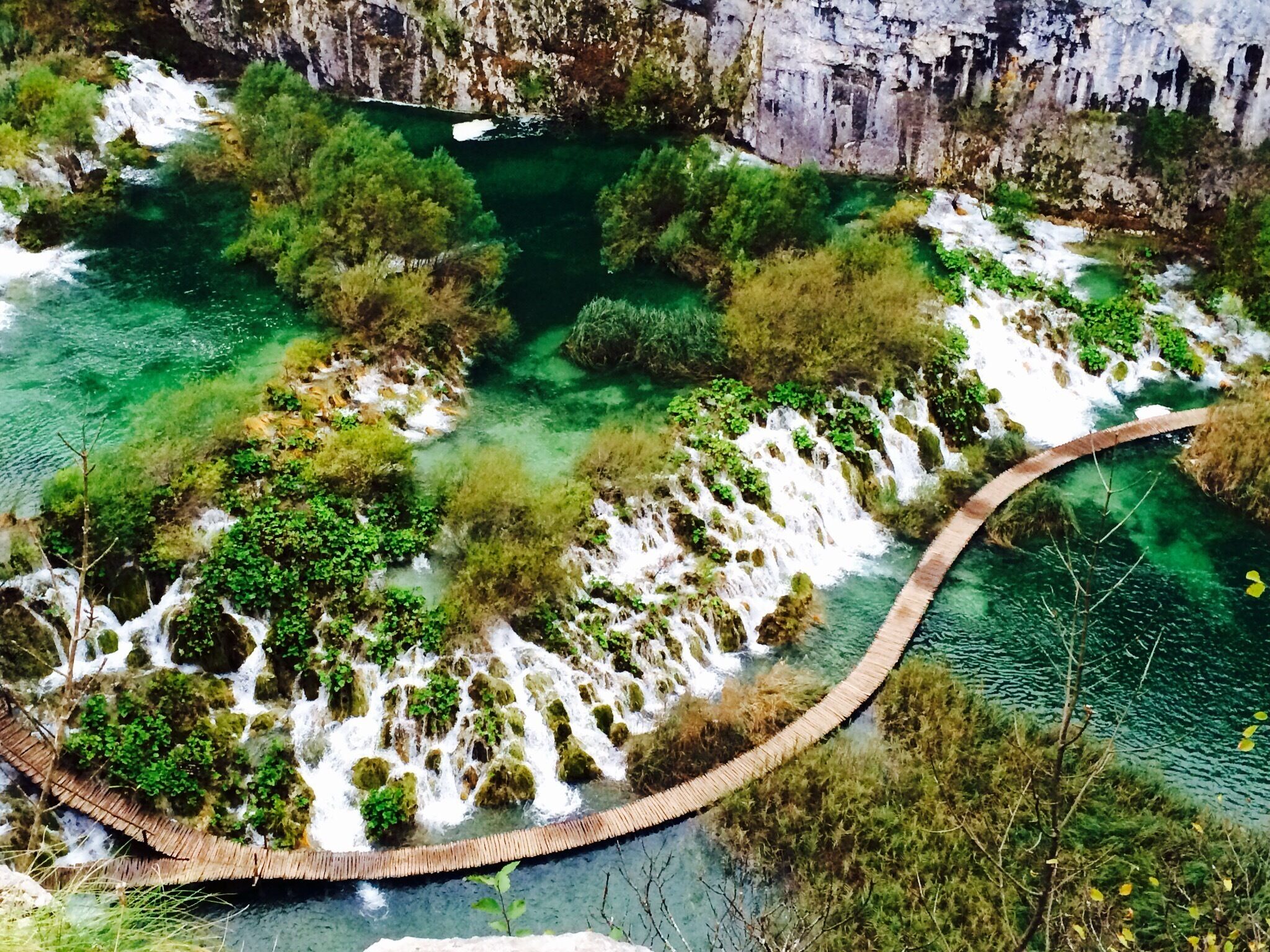 A circular wooden bridge over a clear blue river surrounded by lush greenery and rocky cliffs.
