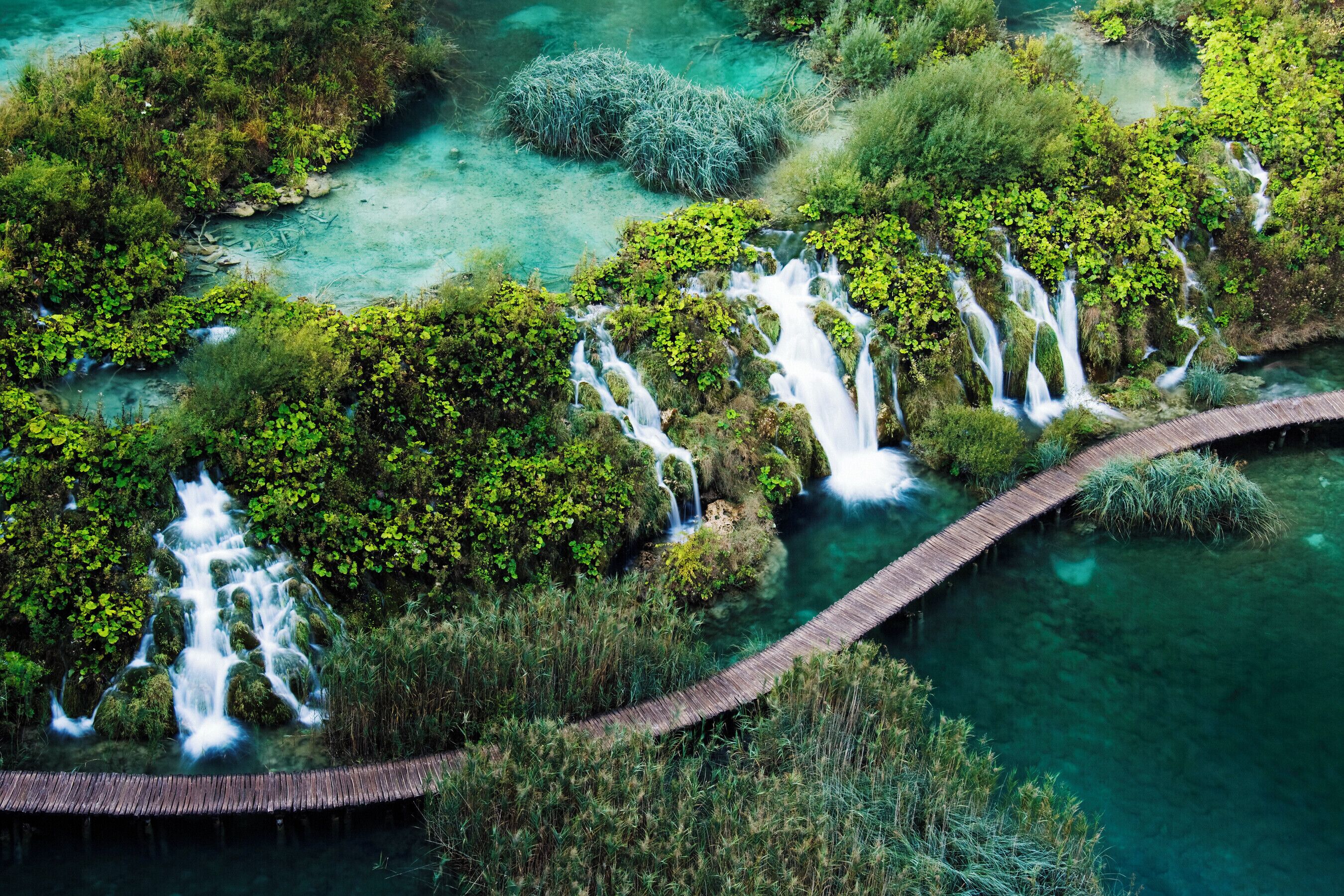 A lush, green landscape with multiple waterfalls and a wooden bridge.