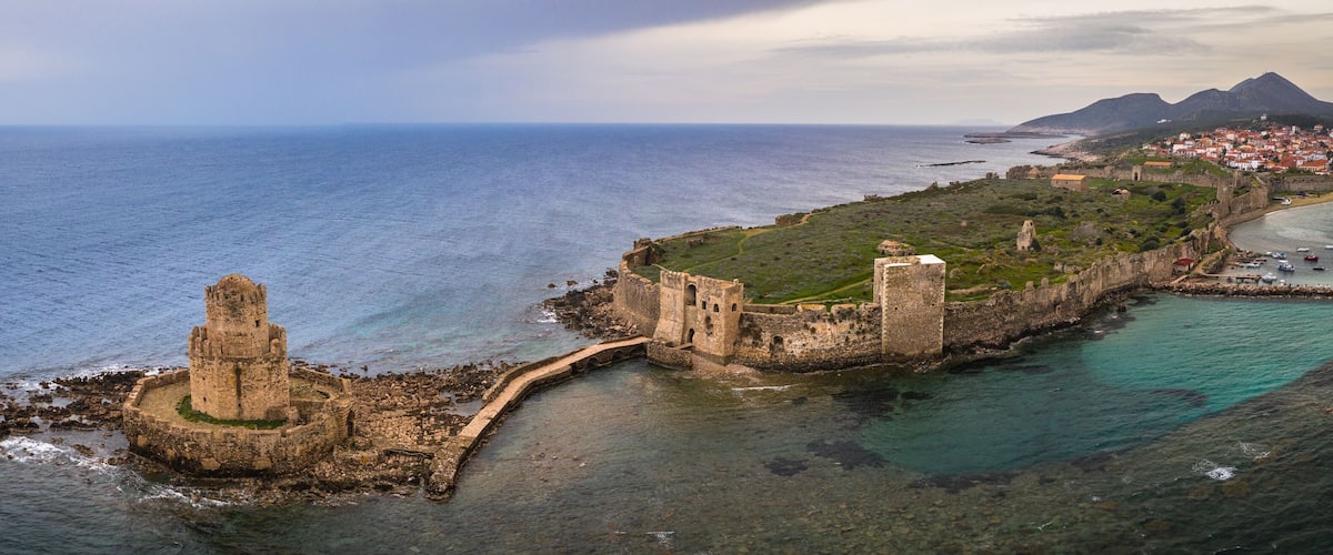 A coastal fortification with a prominent tower, a small town with red-roofed buildings, and a mountainous backdrop.