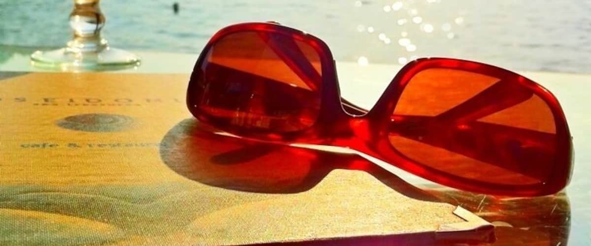 A pair of red sunglasses, a glass of orange drink, and a scenic view of a lake with a distant shoreline.