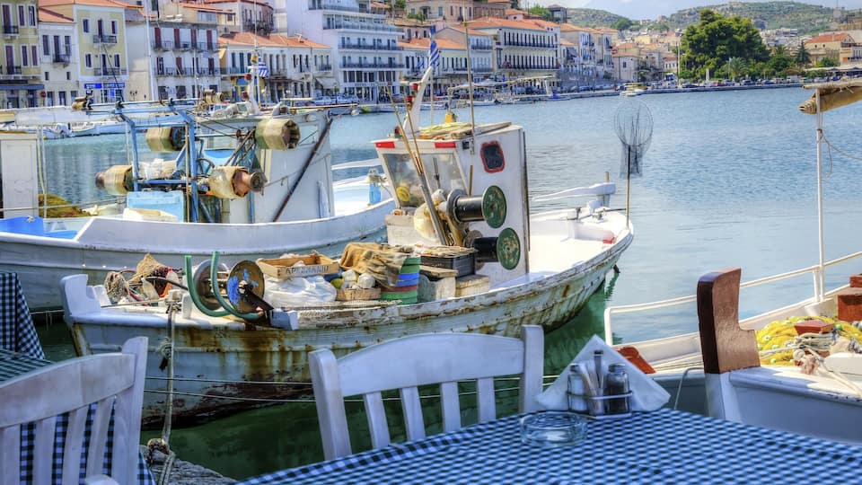 A harbor with boats docked, a table with a checkered tablecloth, and buildings in the background.