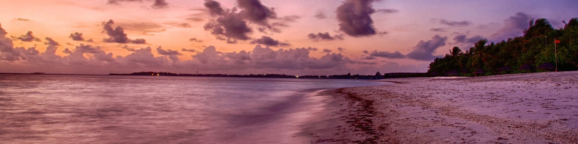 Una playa al atardecer con un mar tranquilo, una orilla arenosa y una costa lejana con árboles y una bandera.