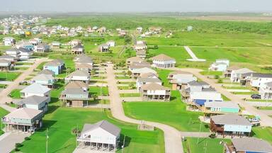 Panorama aerial view long stretch of oceanfront residential development with low-density colorful raised beach houses, grassy backyards, vacant land near sandy shoreline in Port Bolivar, Texas