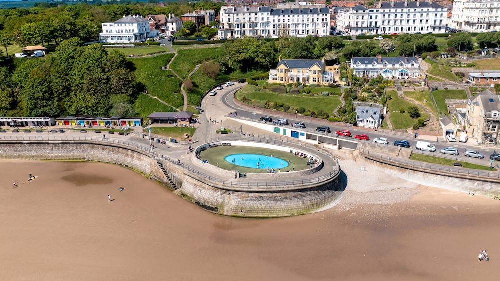 An aerial view of a coastal town with a roundabout, a swimming pool, and residential buildings.