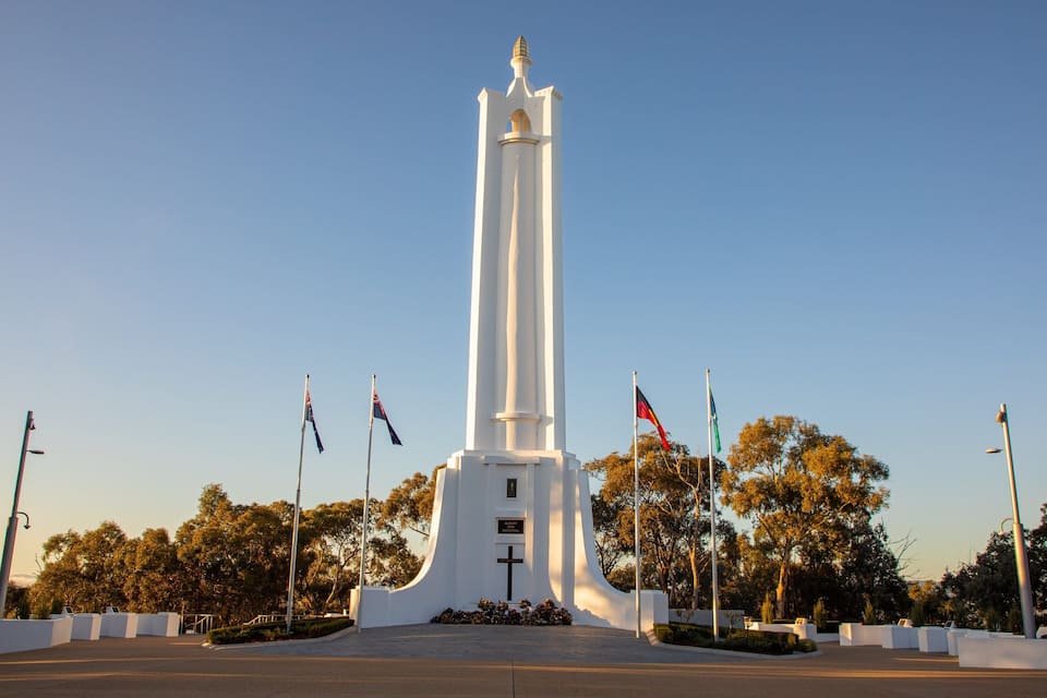 A tall white monument with a cross at the base, surrounded by trees and flags.