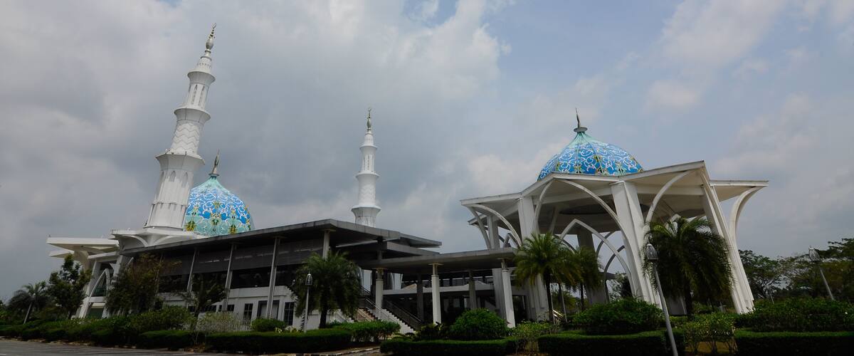 A mosque with two minarets and a blue dome, surrounded by greenery.