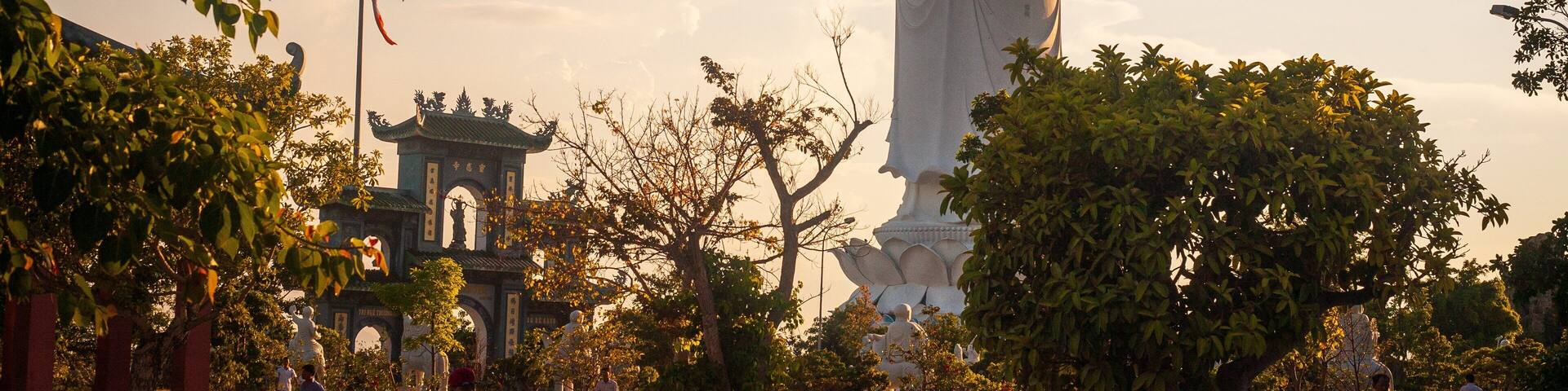 Una gran estatua blanca en un parque con árboles y arquitectura tradicional al fondo.