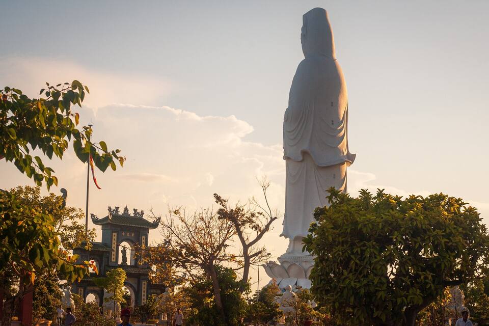 Une grande statue blanche se dresse dans un parc, avec des arbres et des bâtiments à l’architecture traditionnelle en arrière-plan.