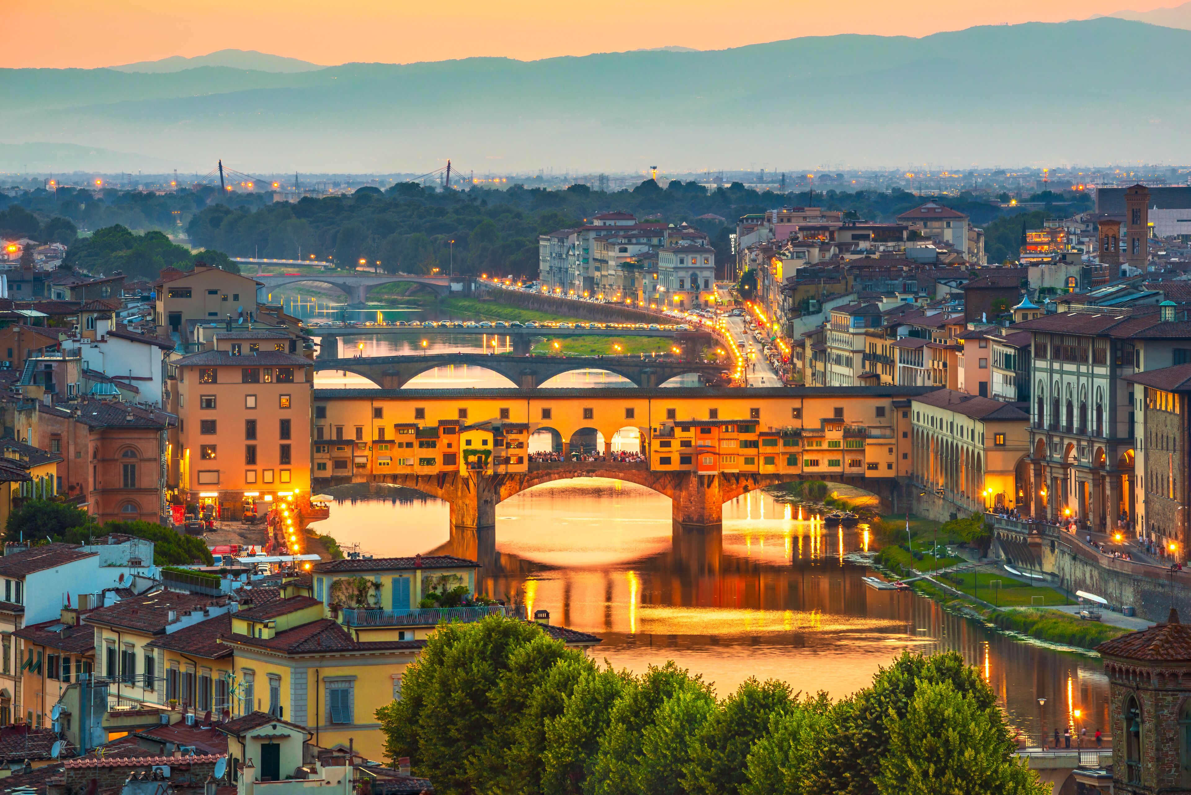 A cityscape with a prominent bridge over a river, surrounded by historic buildings and a distant mountain range.